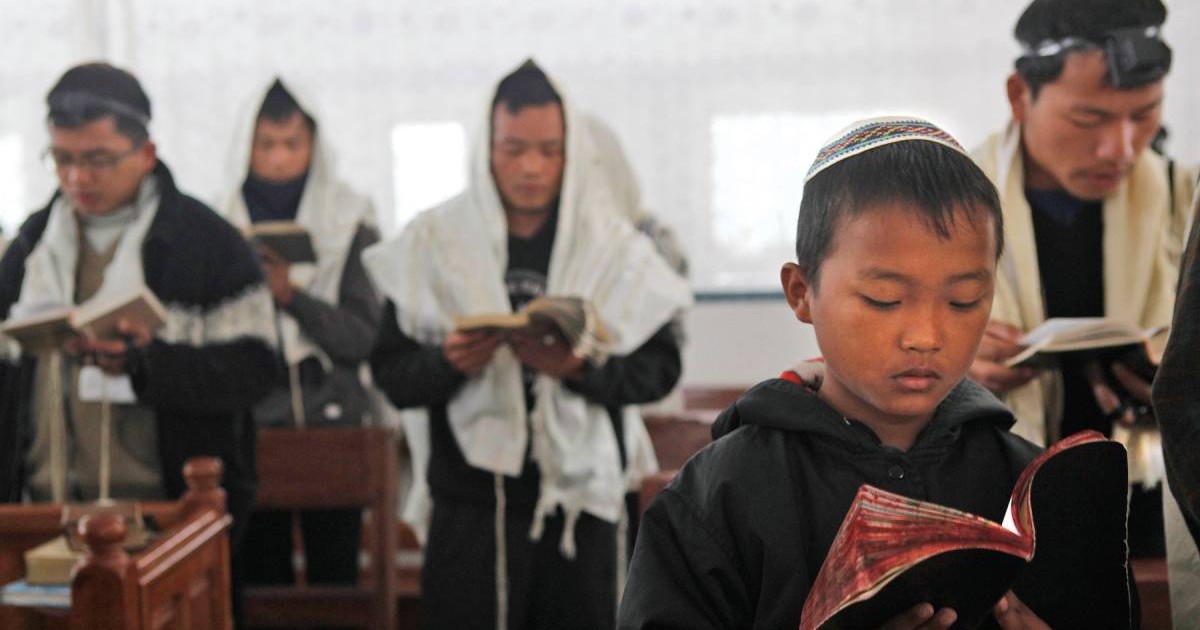 Imaginea arată câțiva membri ai comunității Bnei Menashe din India, aflați la rugăciune. În prim-plan este un copil care citește dintr-o carte, iar în spate se văd bărbați purtând obiecte tradiționale evreiești.