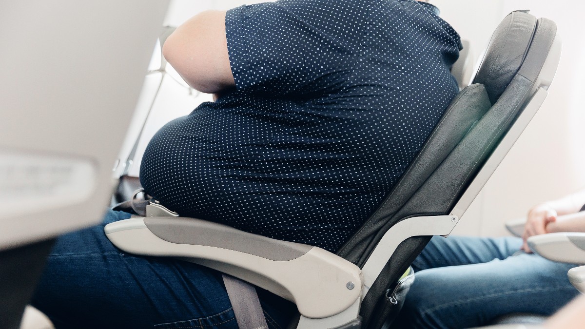 Imaginea prezintă o persoană, văzută din spate, cu un corp voluminos care umple complet scaunul unui avion, sugerând că este supraponderală. Persoana poartă o cămașă neagră cu buline albe și pantaloni din denim închis la culoare, în timp ce mâna sa dreaptă este prinsă în jurul centurii de siguranță.
