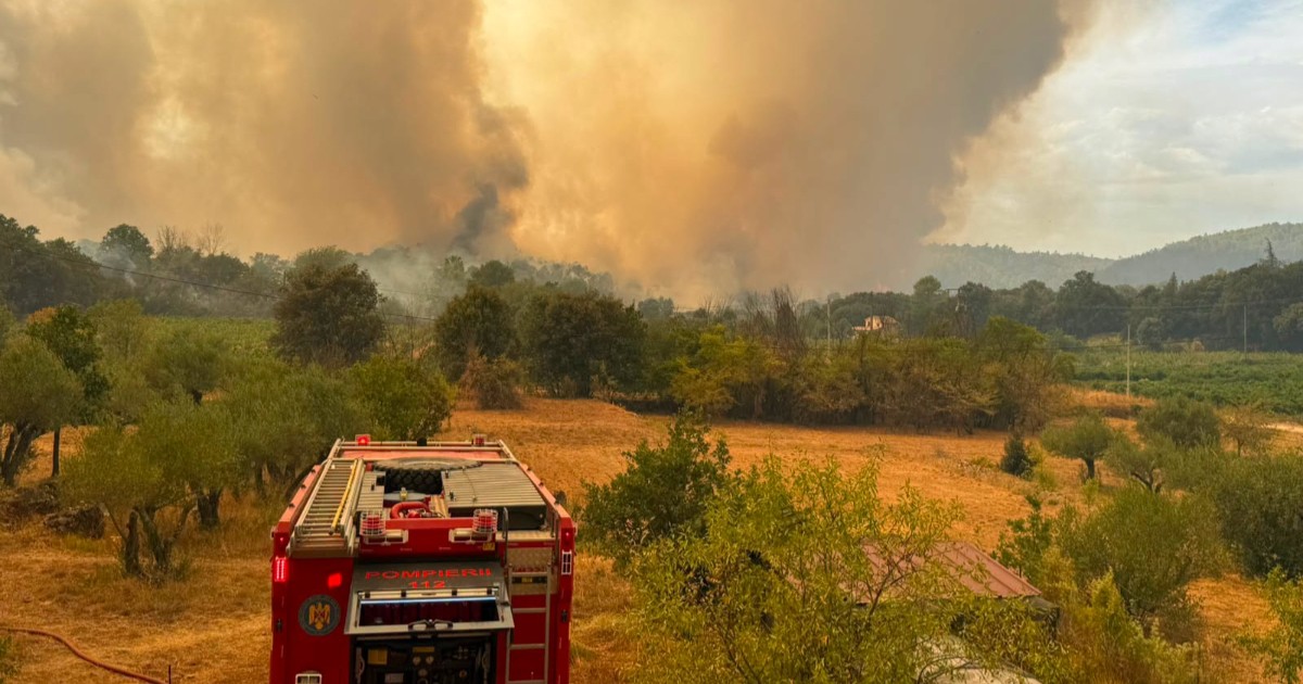 O mașină de pompieri pe un câmp din Grecia, în timpul unui incendiu de vegetație, în fundal se vede cum iese fum