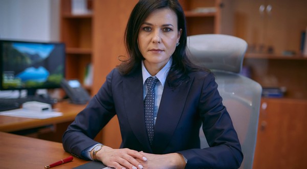 Cristina Chiriac în sacou albastru închis, cămașă albastră și cravată, în timp ce stă la birou