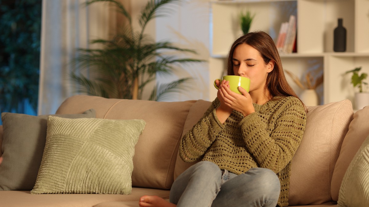 O femeie stă relaxată pe canapea, într-un living confortabil, și bea o băutură caldă dintr-o cană. Atmosfera este liniștită, sugerând un moment de relaxare de seară.