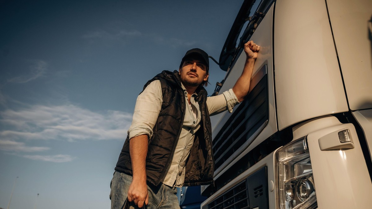 Poză cu un șofer care se sprijină cu o mână de un camion. Are în mână o geantă