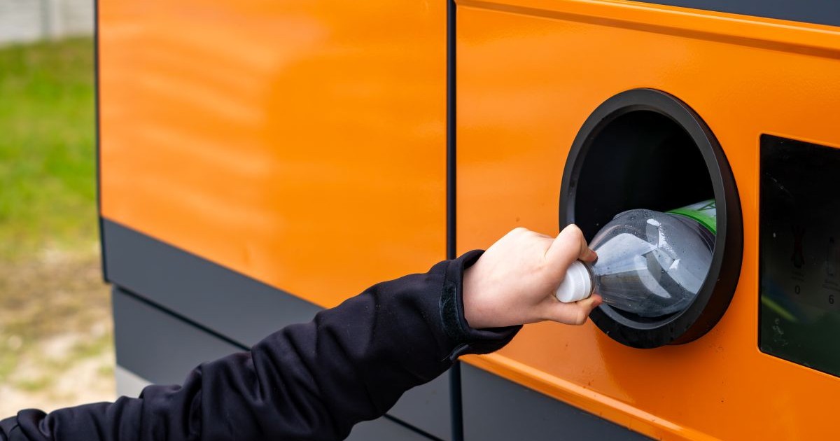 Mâna unui om, îmbrăcat cu o bluză neagră cu mânecă lungă, care introduce o sticlă în aparat portocaliu de reciclat, așezat pe un spațiu verde