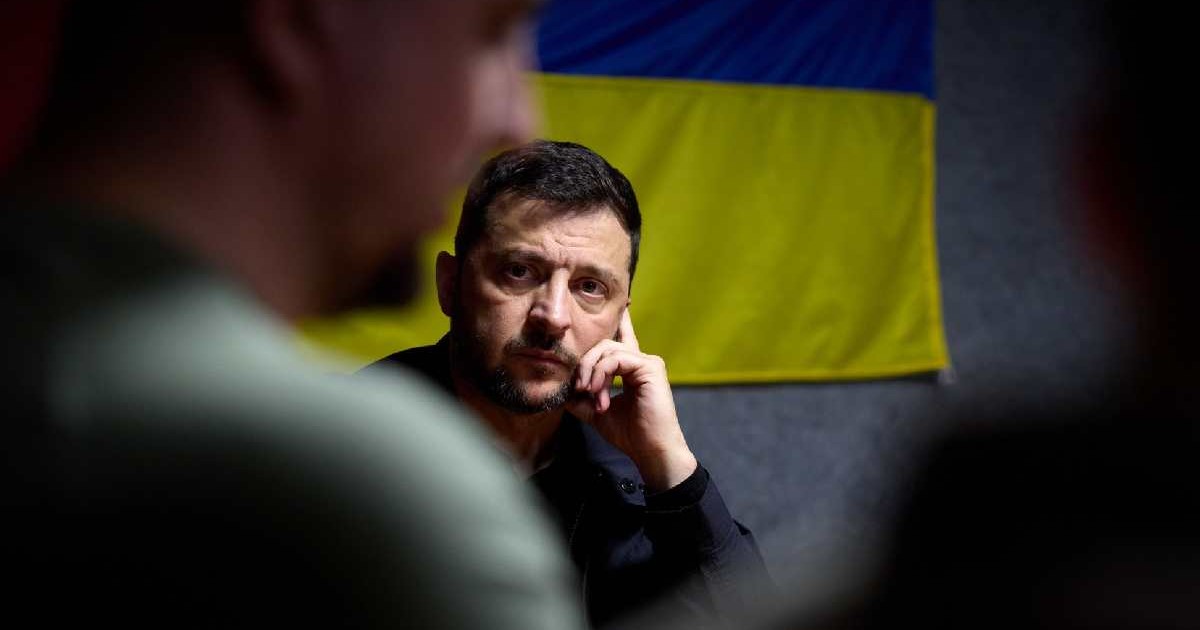 Volodimir Zelenski pe gânduri, cu mâna la față, în timpul unei discuții. În spatele său se află steagul Ucrainei