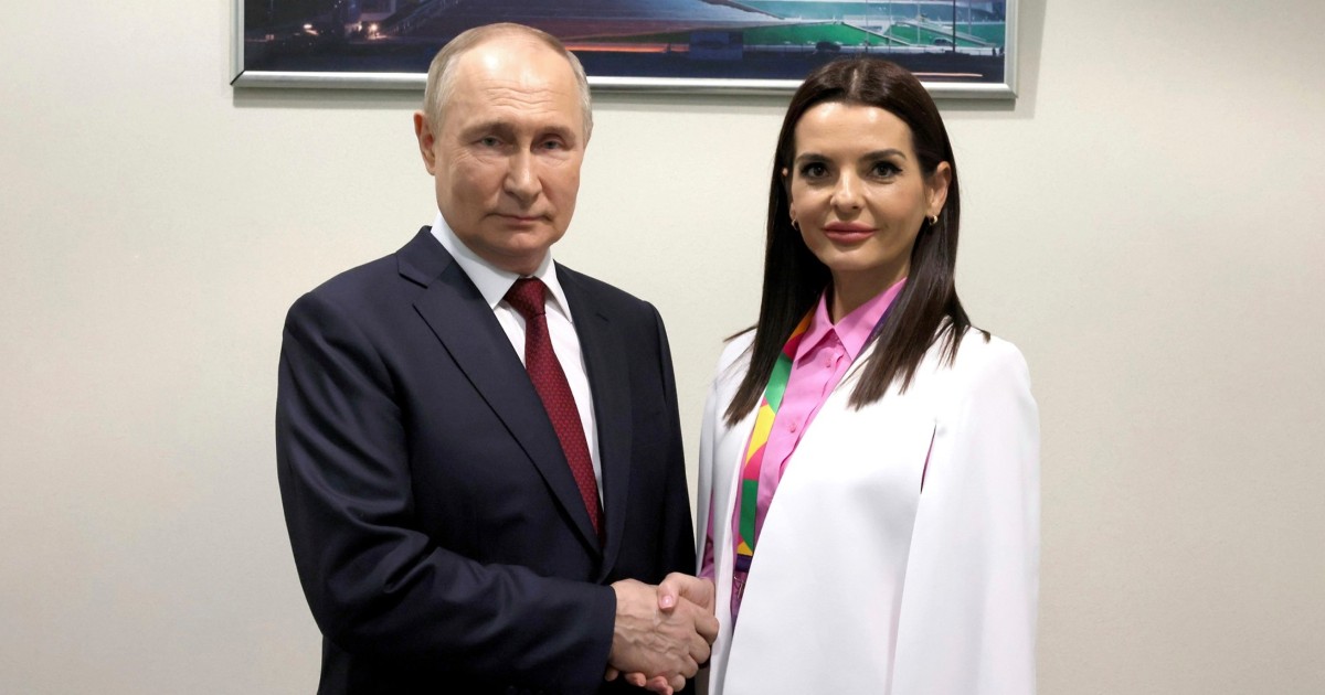 Vladimir Putin, îmbrăcat într-un costum bleumarin, cămașă albă și cravată roșie, dă mâna cu Evghenia Guțul, îmbrăcată cu un sacou alb si cămașă roz. Pe fundal, un perete alb și un tablou în partea de sus.