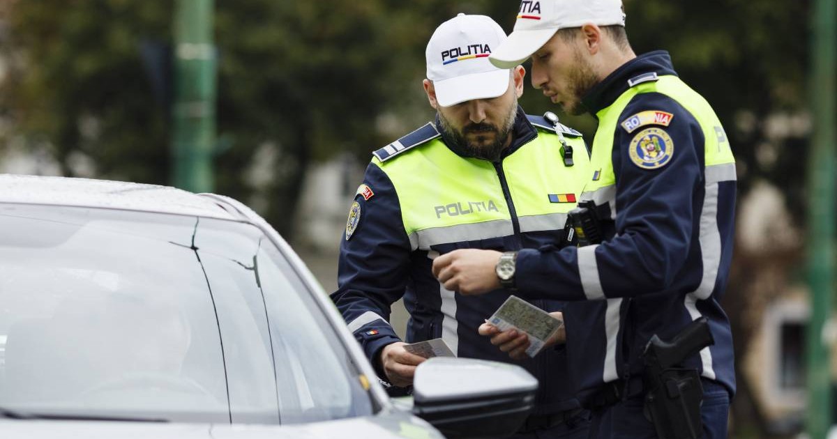 Doi agenți ai Poliției Rutiere din România, îmbrăcați în uniformă. Cei doi verifică documente, cel mai probabil un permis de conducere și un talon auto, în timpul unei opriri în trafic. Aceștia se află lângă un autoturism de culoare argintie.