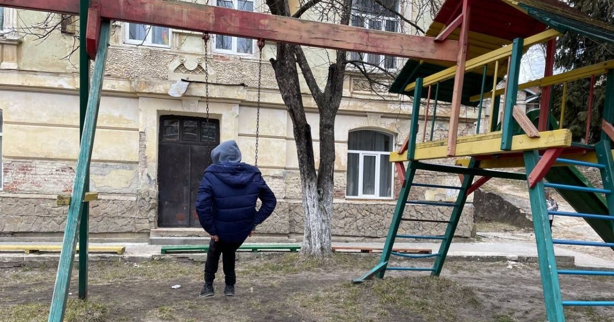 Un băiat cu geacă și glugă pe cap se dă într-un leagăn în fața unui orfelinat din Ucraina, martie 2025