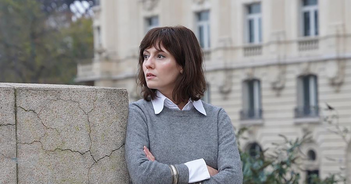Ada Galeș, îmbrăcată cu o bluză de culoare gri care acoperă o cămașă de culoare albă, sprijinită de un perete
