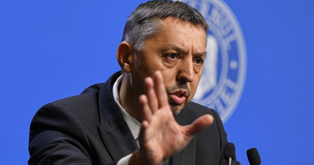 Daniel David, ministrul Educației, îmbrăcat în costum, la pupitrul de la Guvernul României, într-o conferință de presă, cu microfoane în față și logo-ul Guvernului pe fundal albastru în spate