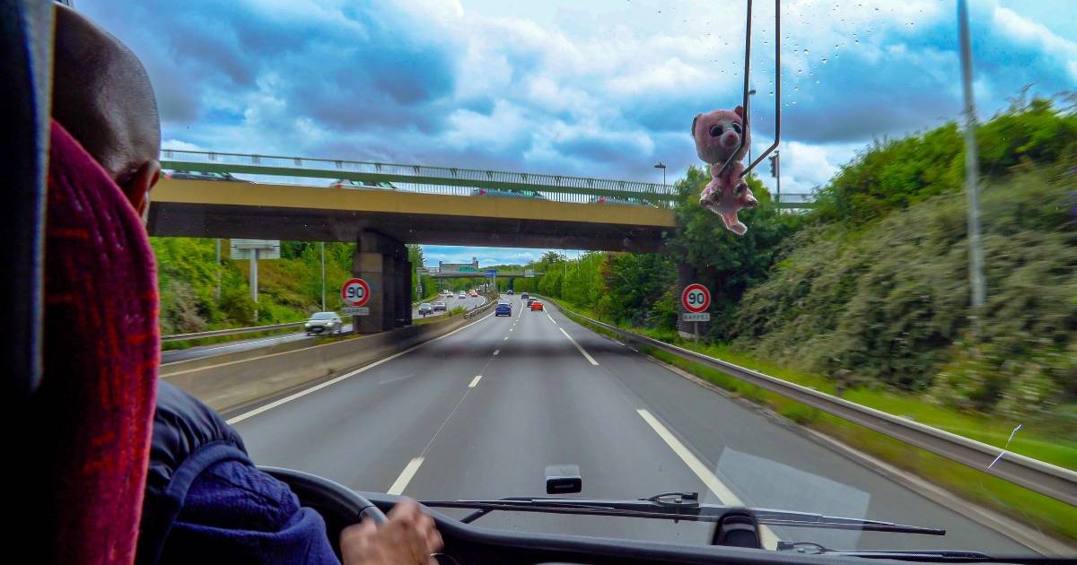 Un șofer tuns scurt, în timp ce conduce un camion pe o autostradă din Franța, ține mâinile pe volan și privește înainte