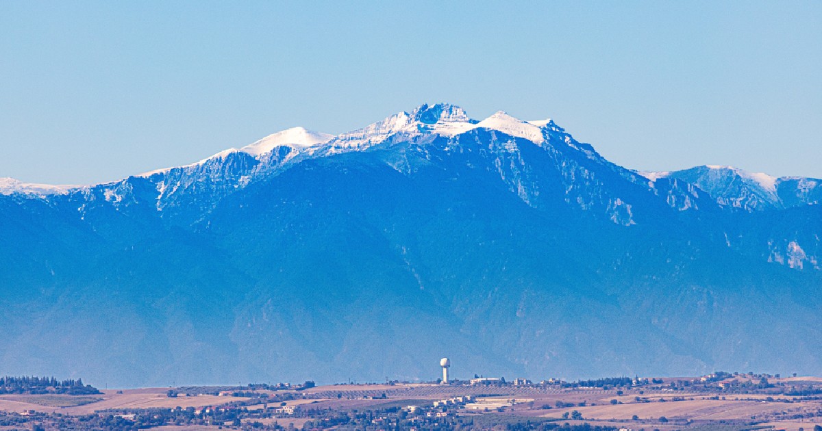 Vedere clară a Muntelui Olimp, cel mai înalt munte din Grecia, cu vârfuri acoperite de zăpadă sub un cer albastru și sate în prim-plan.