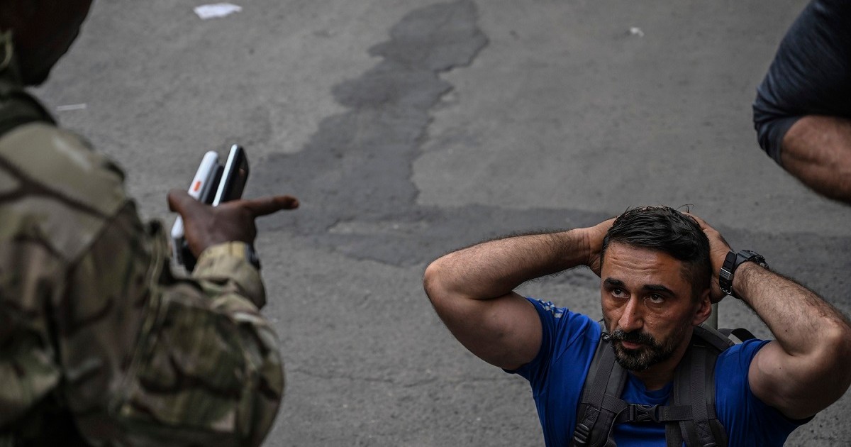 Bărbat în tricou albastru, în genunchi și cu mâinile deasupra capului, certat de un soldat