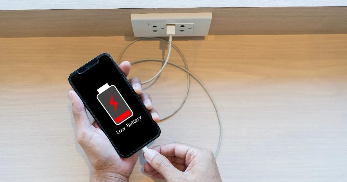 Imaginea arată o persoană care conectează un telefon mobil la încărcător, în timp ce pe ecran apare avertizarea de baterie descărcată. Scena sugerează necesitatea reîncărcării dispozitivului și utilizarea frecventă a telefonului în viața de zi cu zi.