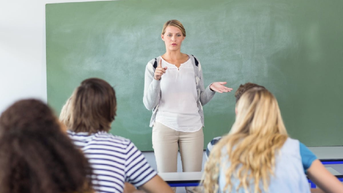 Imaginea prezintă o profesoară stând în fața unei clase de elevi. Ea este îmbrăcată casual, cu o cămașă albă, un cardigan gri deschis și pantaloni bej. Are părul blond, prins, și o expresie facială serioasă, cu o mână întinsă și un deget ridicat, ca și cum ar explica sau accentua un punct important. Cu cealaltă mână, face un gest deschis, sugerând o discuție. În fundal se află o tablă de culoare verde, iar în prim-plan, se văd din spate capetele elevilor, așezați la pupitre. Unii dintre ei au capetele întoarse, ascultând-o pe profesoară. Atmosfera generală a imaginii este de studiu și de predare-învățare, specifică unui mediu școlar.
