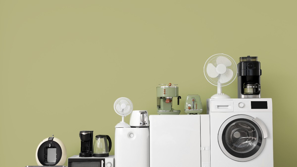 Imaginea prezintă mai multe electrocasnice aliniate într-o locuință, precum mașină de spălat, frigider, cuptor cu microunde, cafetieră și ventilatoare, pe un fundal simplu. Vizualul sugerează consumul zilnic de energie dintr-o gospodărie modernă și diversitatea aparatelor care contribuie la factura de electricitate.