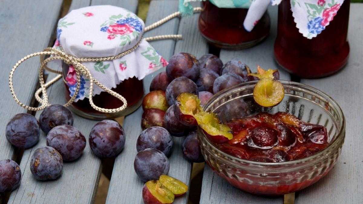 Un bol cu dulceață de prune, prune întregi și borcane de gem pe o masă de lemn
