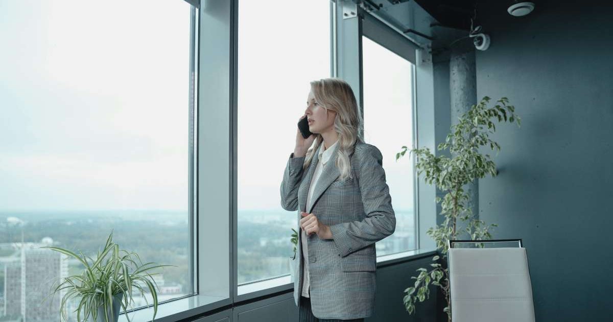 Femeie la birou, imbracata business, vorbeste la telefon si se uita pe geam