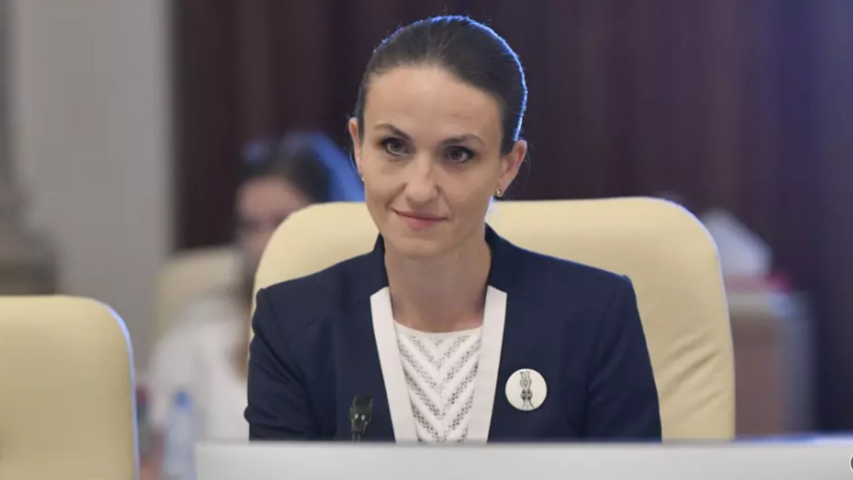 Oana Țoiu îmbrăcată într-un sacou negru și bluză albă în timp ce stă așezată pe un scaun