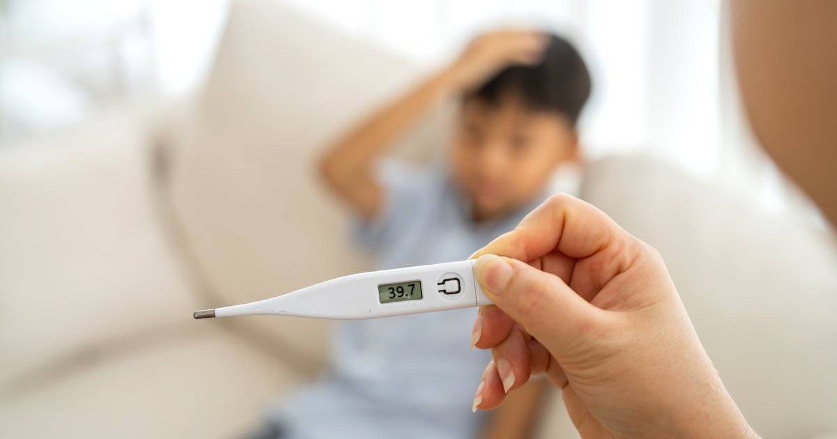 Un copil stând pe canapea, cu o palmă la cap, mama i-a luat temperatura și termometru indică 39