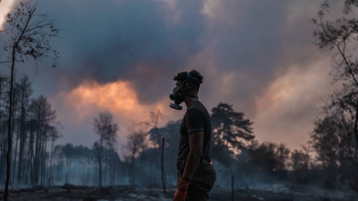 Un bărbat poartă o mască de protecție respiratorie într-o zonă afectată de incendii de vegetație, în timp ce un nor dens de fum acoperă cerul și copacii din jur.