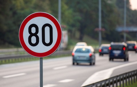 Un indicator de limită de viteză de 80 km/h care se vede în prim plan, iar pe fundal o autostradă, în blur, pe care se văd mașini care circulă