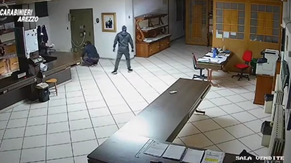 Secvență surprinsă de o cameră de supraveghere dintr-o încăpere, în care două persoane mascate și îmbrăcate în haine închise la culoare încearcă să spargă un seif metalic montat pe un perete.