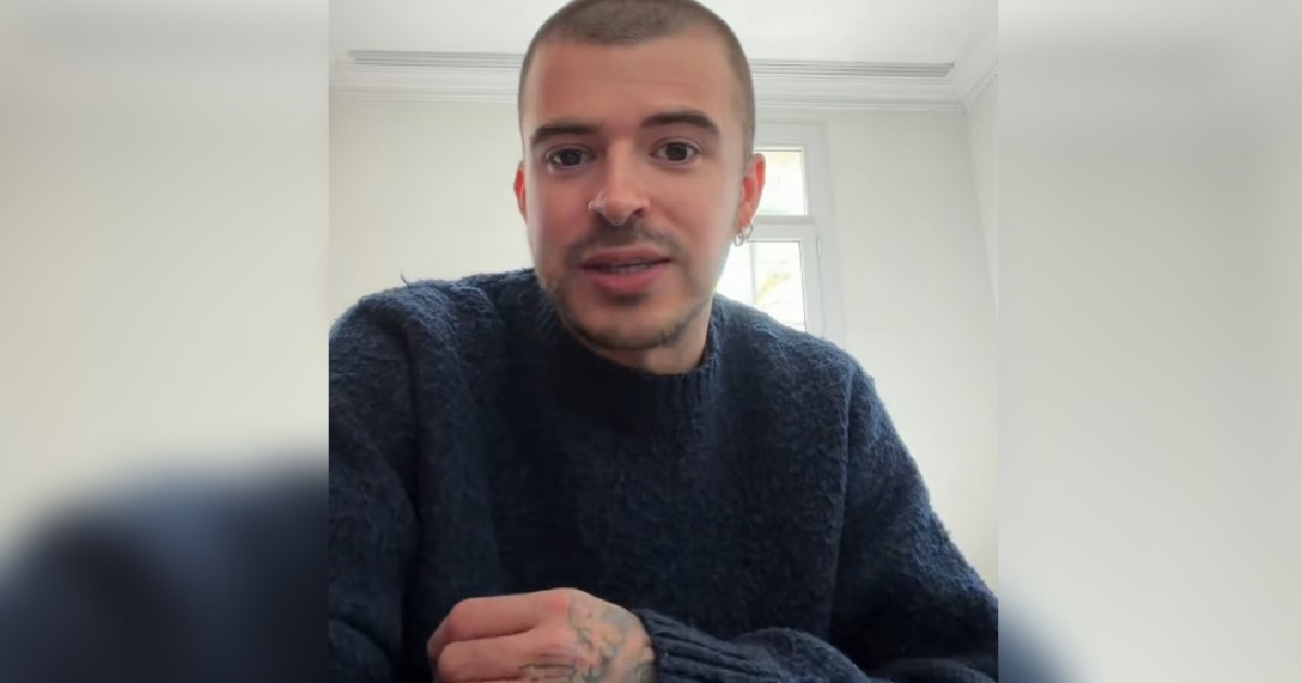 Imaginea surprinde un tânăr cu părul tuns foarte scurt și mâna tatuată, purtând un pulover albastru închis, în timp ce pare să vorbească în fața unei camere într-o încăpere luminoasă.