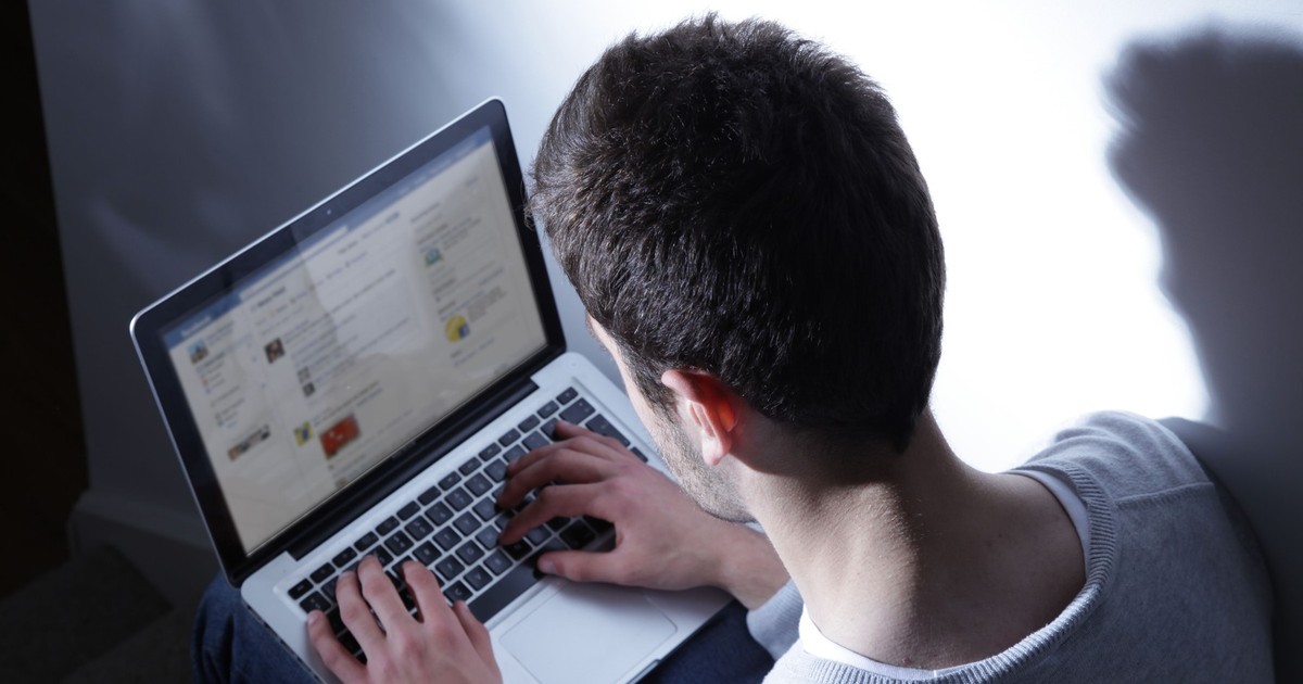 Bărbat surprins în timp ce ține pe picioare un laptop, pe ecranul căruia este afișată platforma Facebook