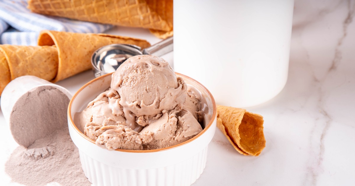 Imaginea prezintă un bol cu mai multe cupe de înghețată cu aromă de ciocolată, alături de cornete crocante și pudră de cacao. Desertul este așezat pe o suprafață deschisă la culoare, într-o compoziție luminoasă și apetisantă.
