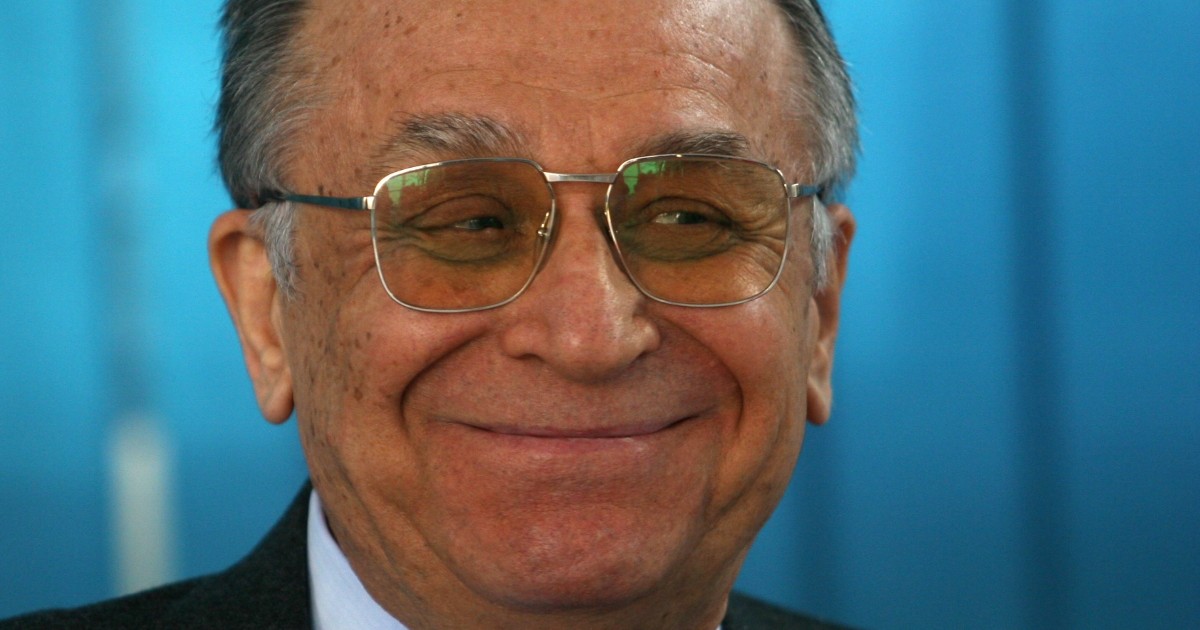 Ion Iliescu, cu ochelari de vedere la ochi, zâmbește, iar în spate are un fundal bleu