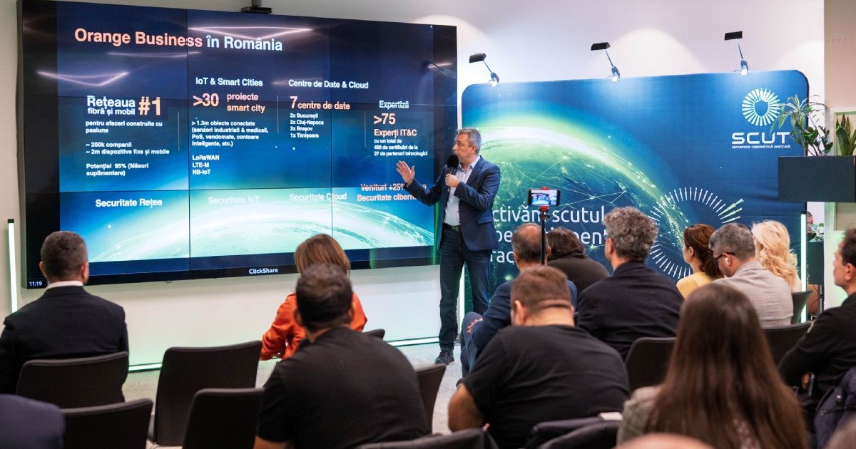 Conferința SCUT și Orange Cyberdefense, cu un bărbat îmbrăcat în costum, care ține un microfon în mână și prezintă date pe panouri digitale, în fața unor oameni așezați pe scaune