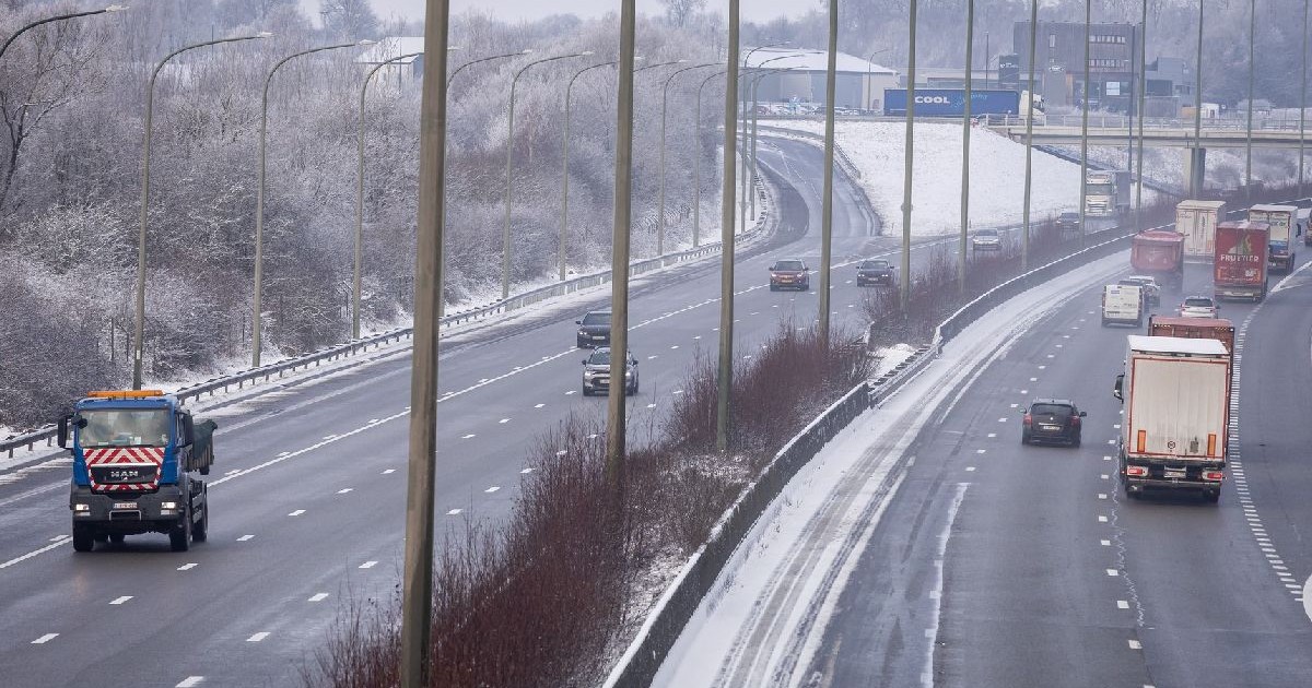O autostradă înzăpezită, cu mai multe benzi, pe care circulă mai multe vehicule, inclusiv un camion albastru și un camion roșu, în ambele sensuri. Copacii și parapetele acoperite de zăpadă mărginesc drumul, iar în fundal se vede un pod.