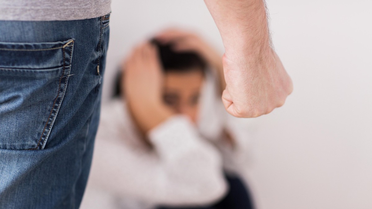 Imagine cu o femeie care stă pe podea și-și ține mâinile pe cap, iar în stânga se află un bărbat cu pumnul încleștat