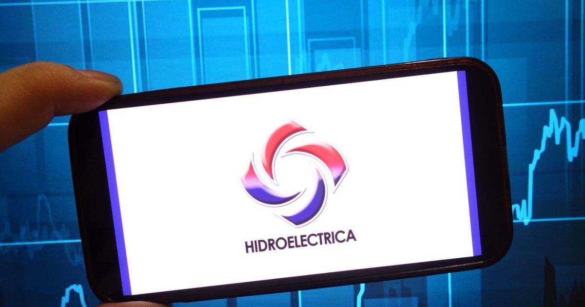 Un om ține în mână un telefon, pe ecranul căruia se vede sigla și numele Hidroelectrica, pe un fundal al cotațiilor bursiere