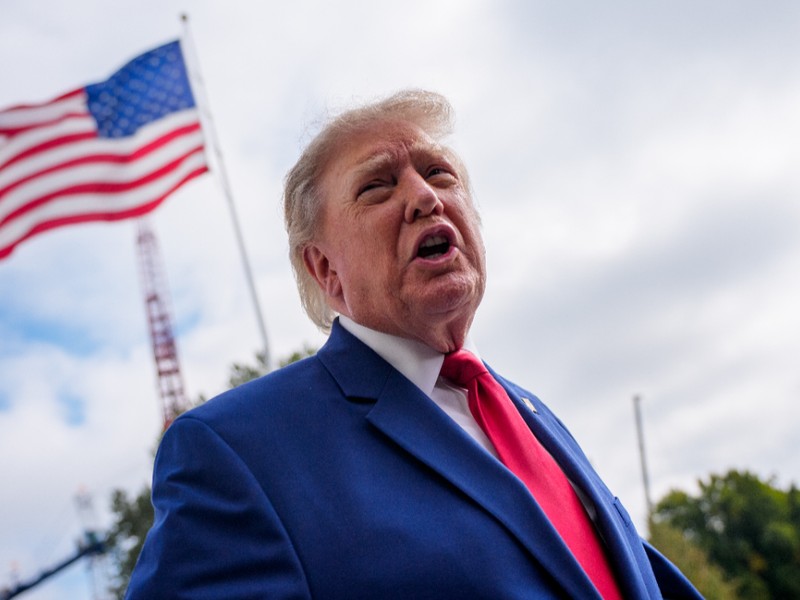 Donald Trump vorbind dintr-un unghi coborât, îmbrăcat în costum albastru și cravată roșie, pe fundalul unui steag al Statelor Unite care flutură în aer liber.