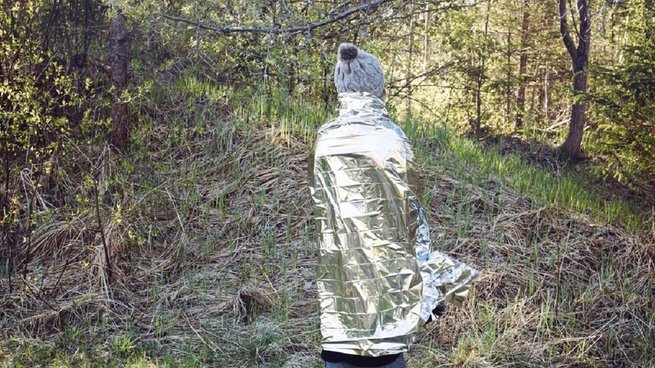 Imagine cu o femeie care s-a rătăcit și se află într-o pădure sălbatică, într-o zi rece, folosind o folie izolantă pentru a preveni hipotermia și pierderea căldurii corporale.