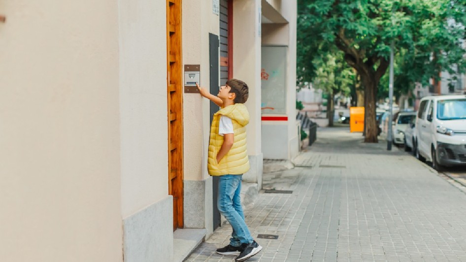 Un băiețel îmbrăcat cu o vestă galbenă și blugi stă pe un trotuar din oraș, întinzându-se pentru a apăsa soneria de la intrarea într-o clădire. În fundal, strada este mărginită de copaci și mașini parcate