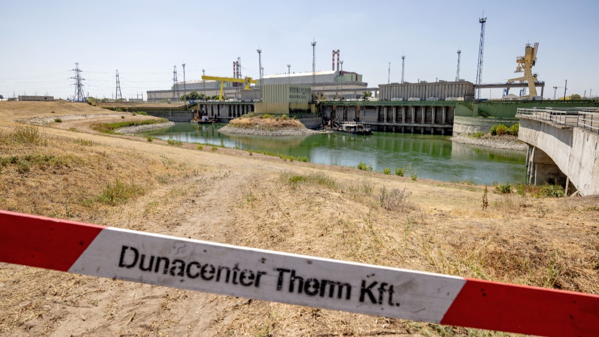 Imaginea prezintă un complex industrial situat lângă un canal de apă, delimitat în prim-plan de o bandă de siguranță alb-roșie pe care este scris „Dunacenter Therm Kft.”.