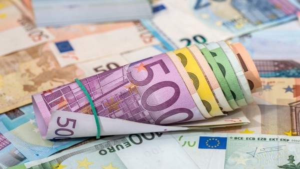 Un sul de bancnote euro de diferite cupiuri, prins cu un elastic verde, așezat peste alte bancnote euro împrăștiate.