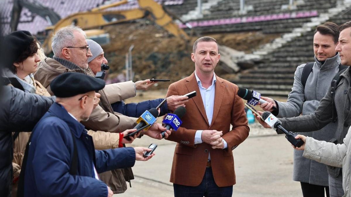 Alfred Simonis surprins în timp ce vorbește în fața jurnaliștilor, așezați și în stânga și în dreapta.