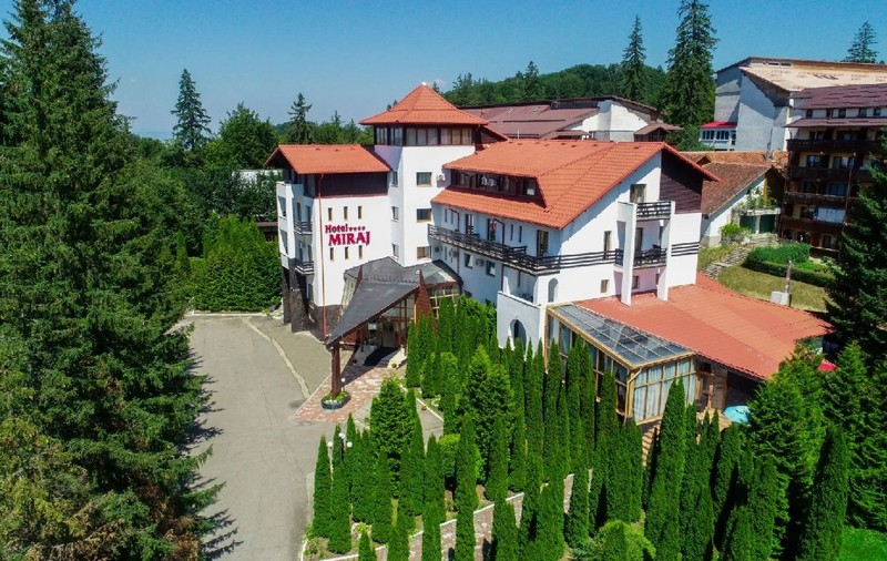 Hotelul Miraj din Poiana Brașov, cladire alba, cu acoperișuri de țiglă roșie, și curte 