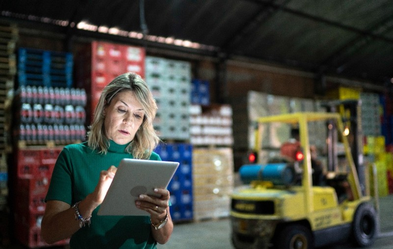O femeie îmbrăcată într-o bluză verde, utilizând o tabletă digitală într-un depozit industrial cu mărfuri stivuite și un motostivuitor galben în fundal.