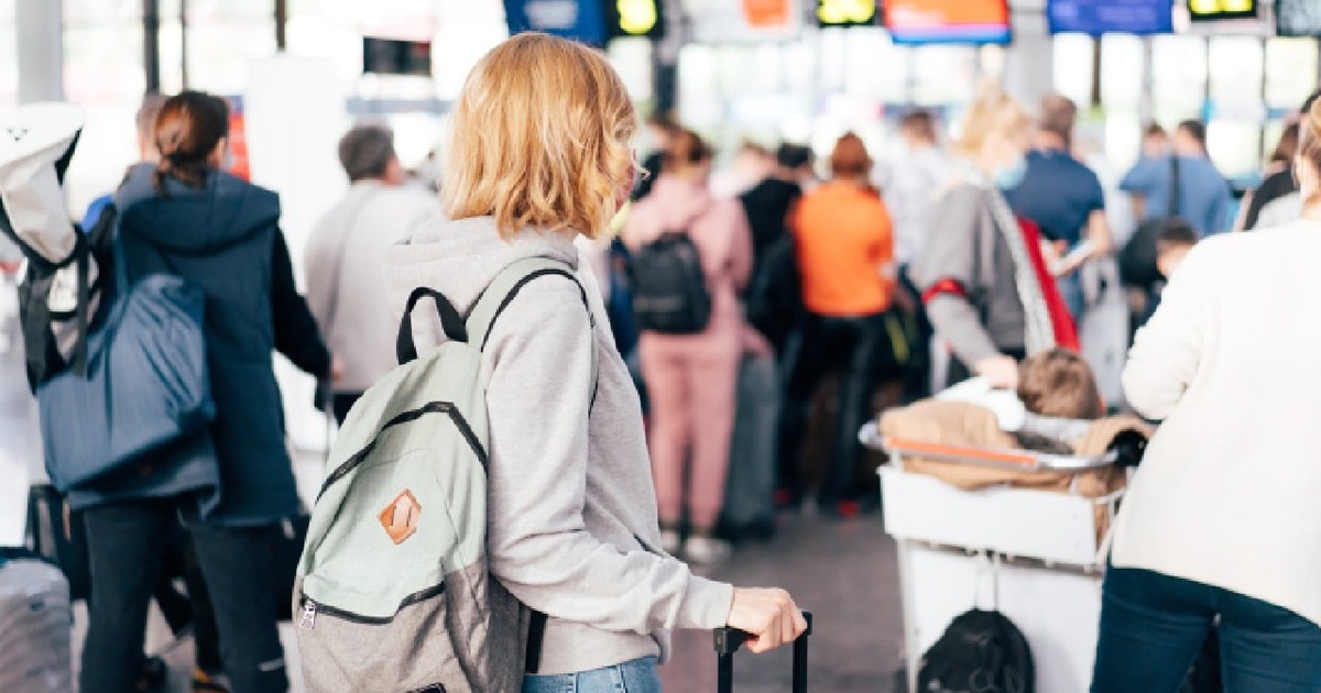 O tânără femeie adultă, cu părul blond și ondulat, stă cu valiza într-un terminal aglomerat. Poartă un hanorac gri, blugi și un rucsac.