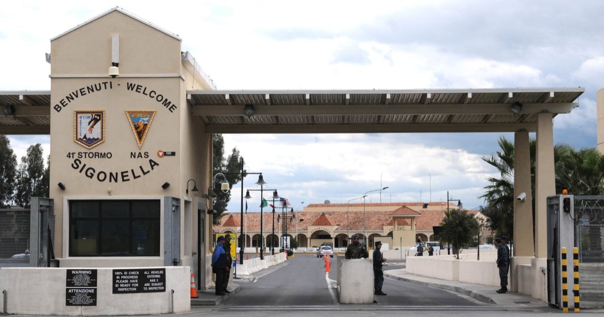 Poarta principală de acces a bazei militare Naval Air Station (NAS) Sigonella, situată în Sicilia, Italia. În partea stângă se află o clădire bej, tip turn, pe care este scris mare: „BENVENUTI - WELCOME” și „41 STORMO - NAS SIGONELLA”.
