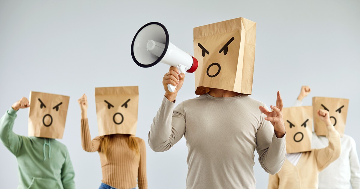 Bărbat cu emoticon furios pe cap strigă tare în difuzor alături de un grup de sprijin. Un grup de oameni cu pungi de hârtie cu emoticoane desenate pe ele scandează și cer acțiuni decisive