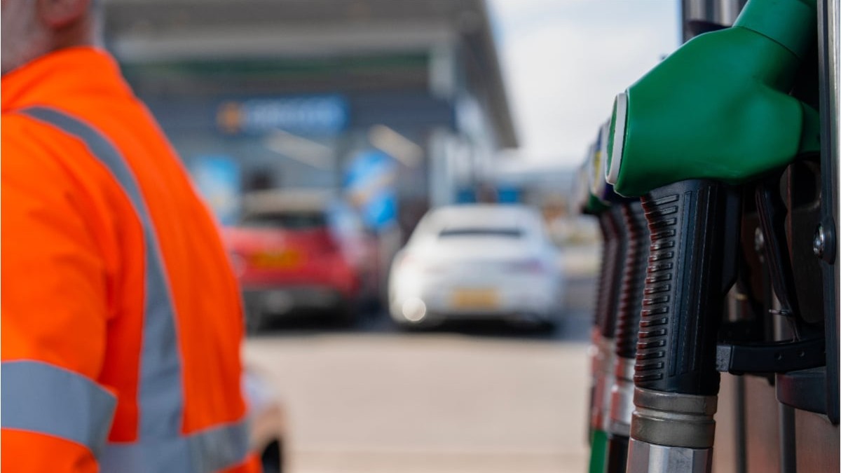 O stație de alimentare cu motorină. Lângă aceasta se află un bărbat îmbrăcat în echipament portocaliu fosforescent.