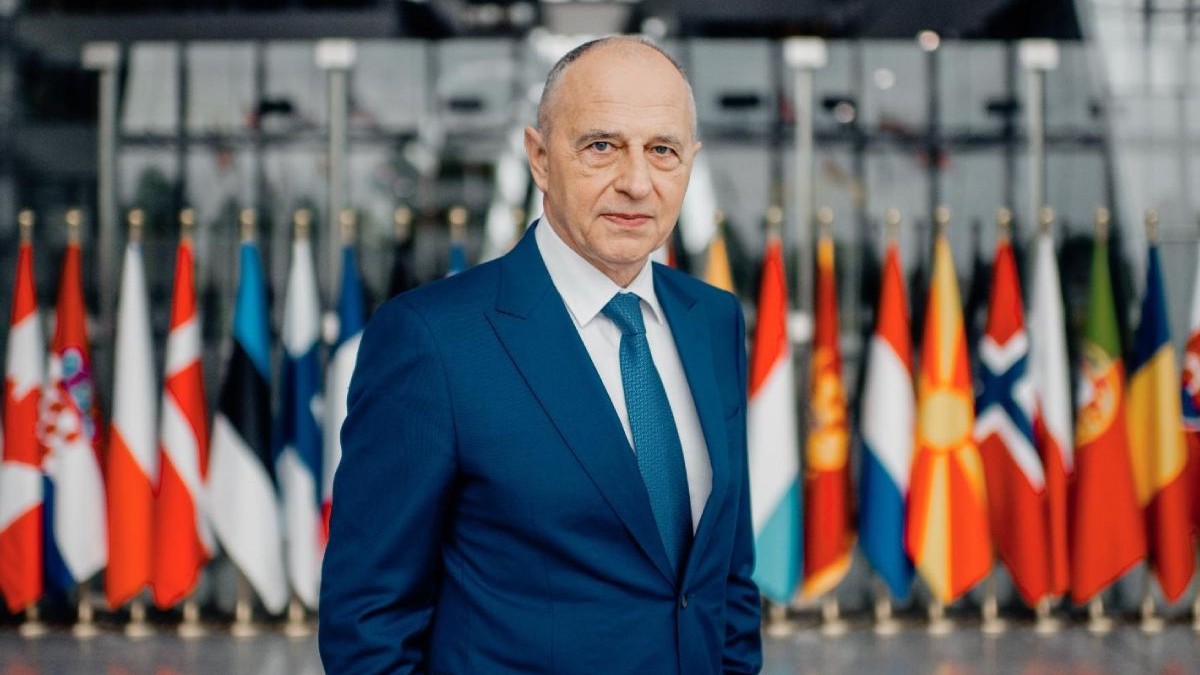 Mircea Geoană, îmbrăcat într-un costum albastru, în picioare, având pe fundal steagurile țărilor NATO