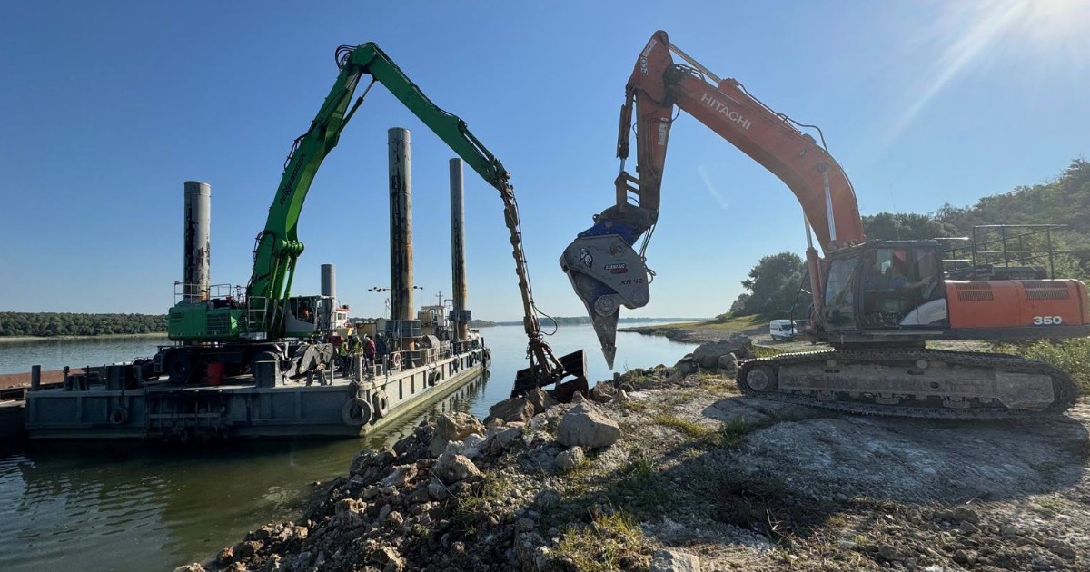 Un excavator care sapă și încarcă pietriș de pe malul Dunării pe o barjă