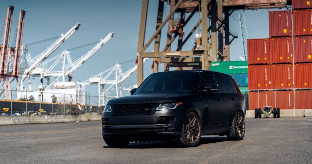 Range Rover de culoare neagră, într-un port, cu containere maro pe fundal și macarale albe și galbene