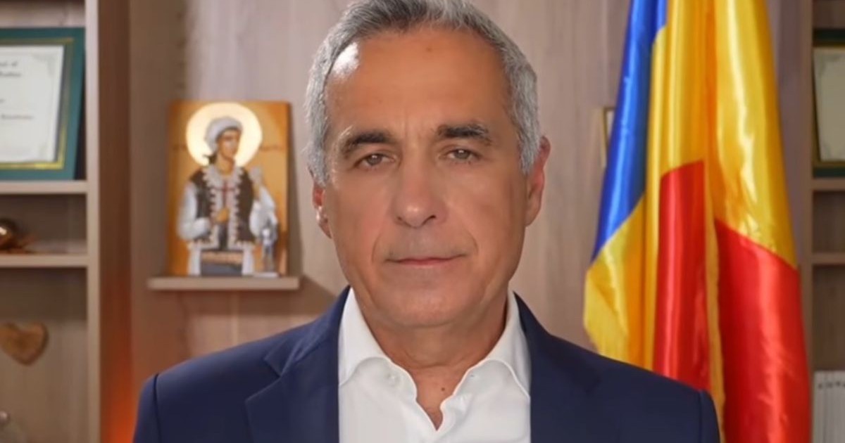 Prim plan Călin Georgescu in sacou negru si camasa albă, cu o bibliotesca in spate, unde se vad o icoană in stanga si un steag al României în dreapta